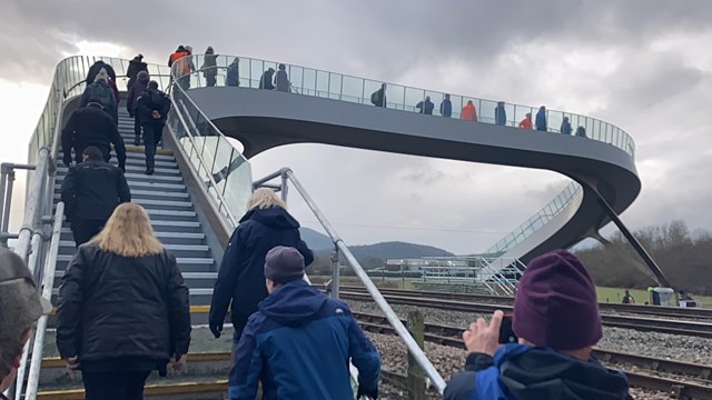 Pedestrians crossing an unusual shaped footbridge over rail tracks