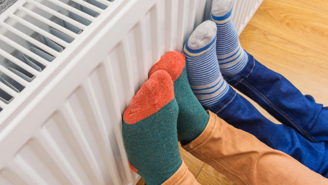 Woman and child wearing colorful pair of woolly socks warming cold feet in front of heating radiator in winter time. Electric or gas heater at home.
