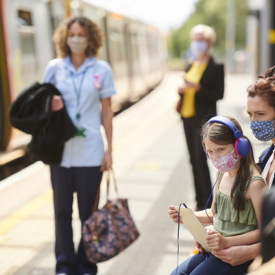 Image of girl and mother sitting on train platform in masks