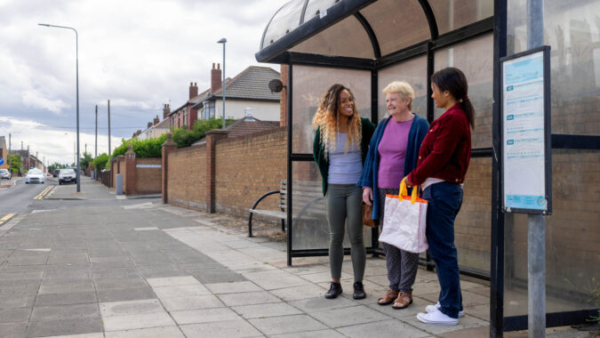 Family members standing, waiting at a bus stop smiling and laughing with each other. They are all wearing casual clothing.