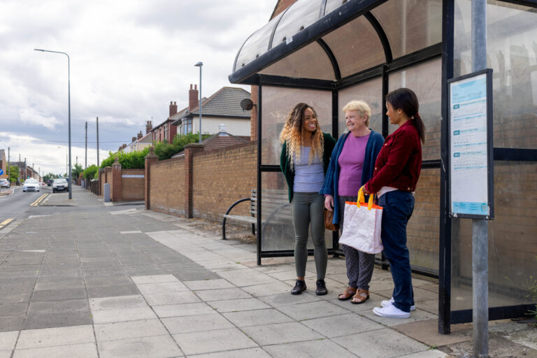 Family members standing, waiting at a bus stop smiling and laughing with each other. They are all wearing casual clothing.