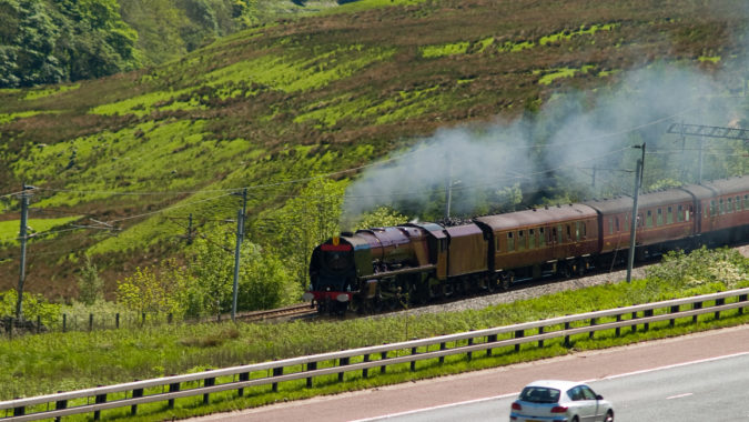 A Princess Coronation (Duchess) class express steam locomotive steaming fast up the west coast main line on the fringes of the English Lake District. A car travelling in the opposite direction is on the adjacent M6 motorway.