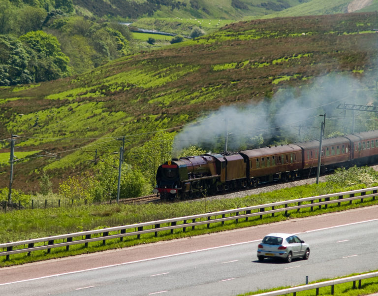 A Princess Coronation (Duchess) class express steam locomotive steaming fast up the west coast main line on the fringes of the English Lake District. A car travelling in the opposite direction is on the adjacent M6 motorway.