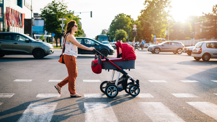 Millennial generation mother with baby in modern red pram walking in downtown on sunny summer day.